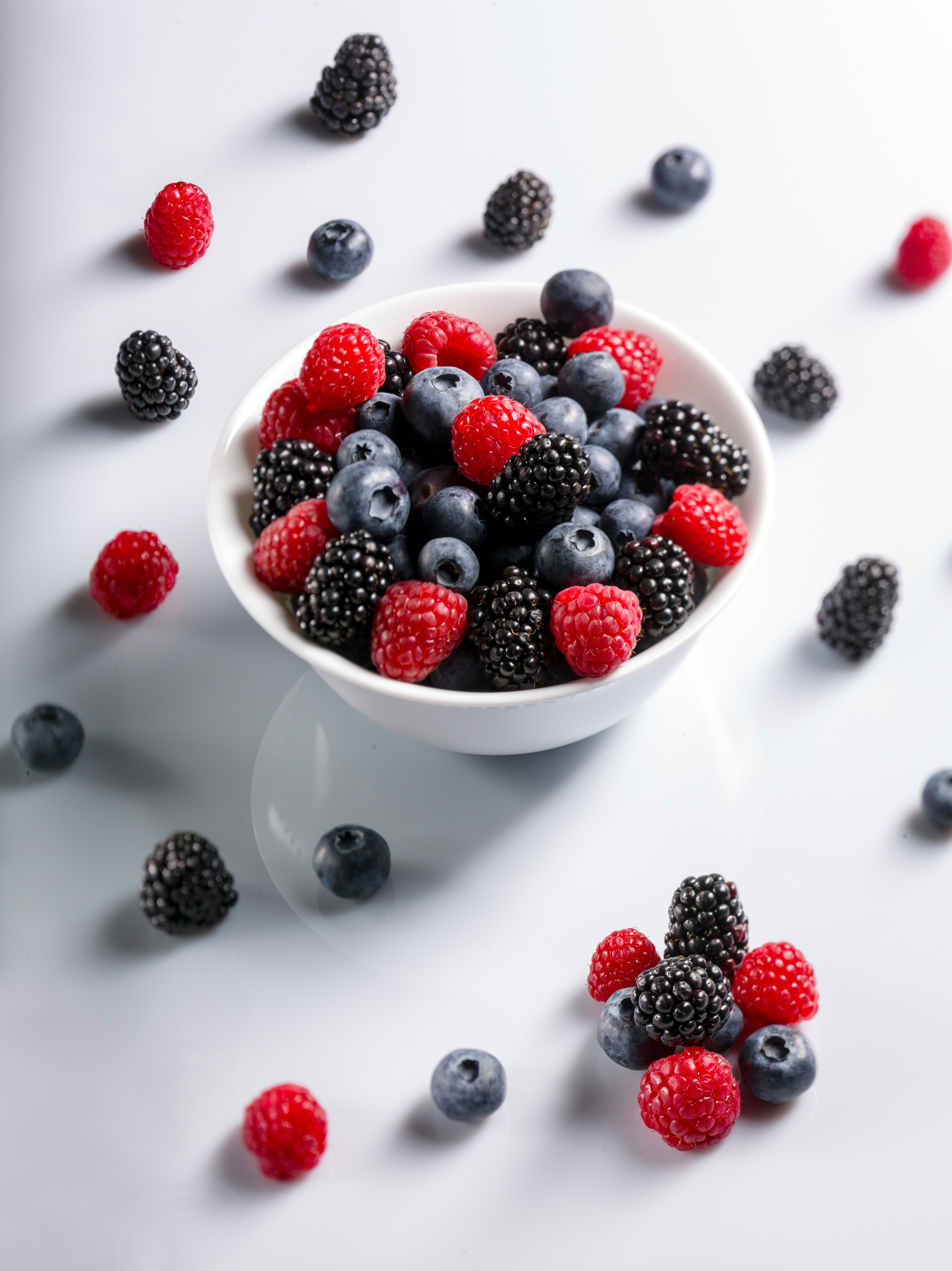 White bowl filled with fresh blueberries, raspberries, and blackberries on a white surface with scattered berries around.